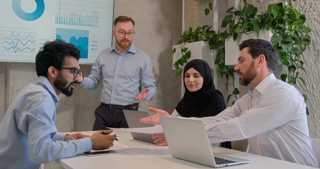Four colleagues in a modern office having a discussion around a white table with laptops and documents, charts displayed on a screen behind them.