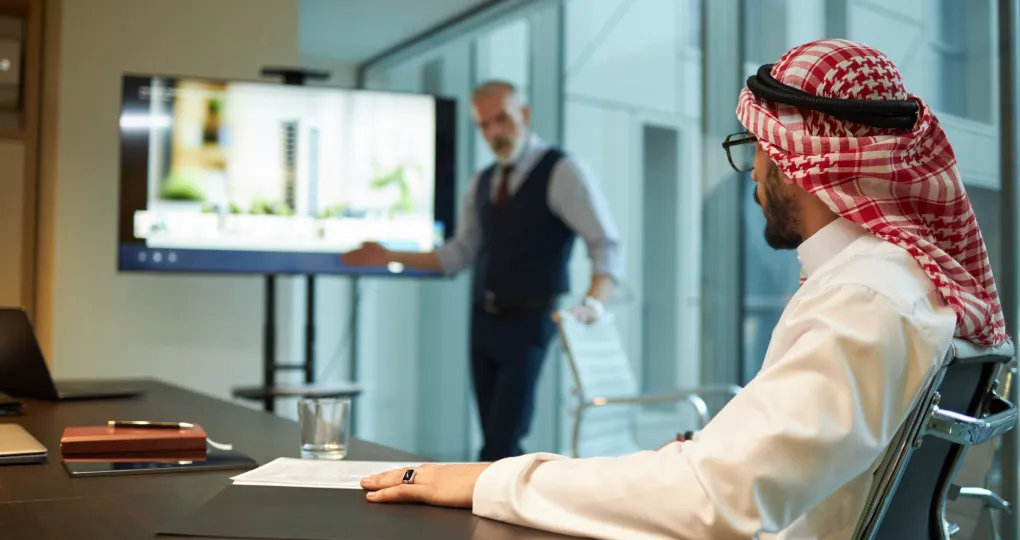 Business meeting in a modern office where a man in traditional Middle Eastern attire listens while a man in a vest presents information displayed on a screen.