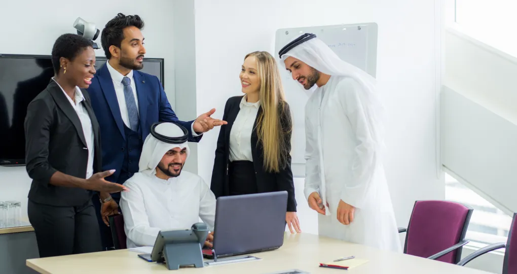 A diverse group of five business professionals, including two men in traditional Middle Eastern attire, a woman in a black blazer, a man in a blue suit, and a woman in a black suit, are gathered around a laptop in a bright office, engaged in a discussion.