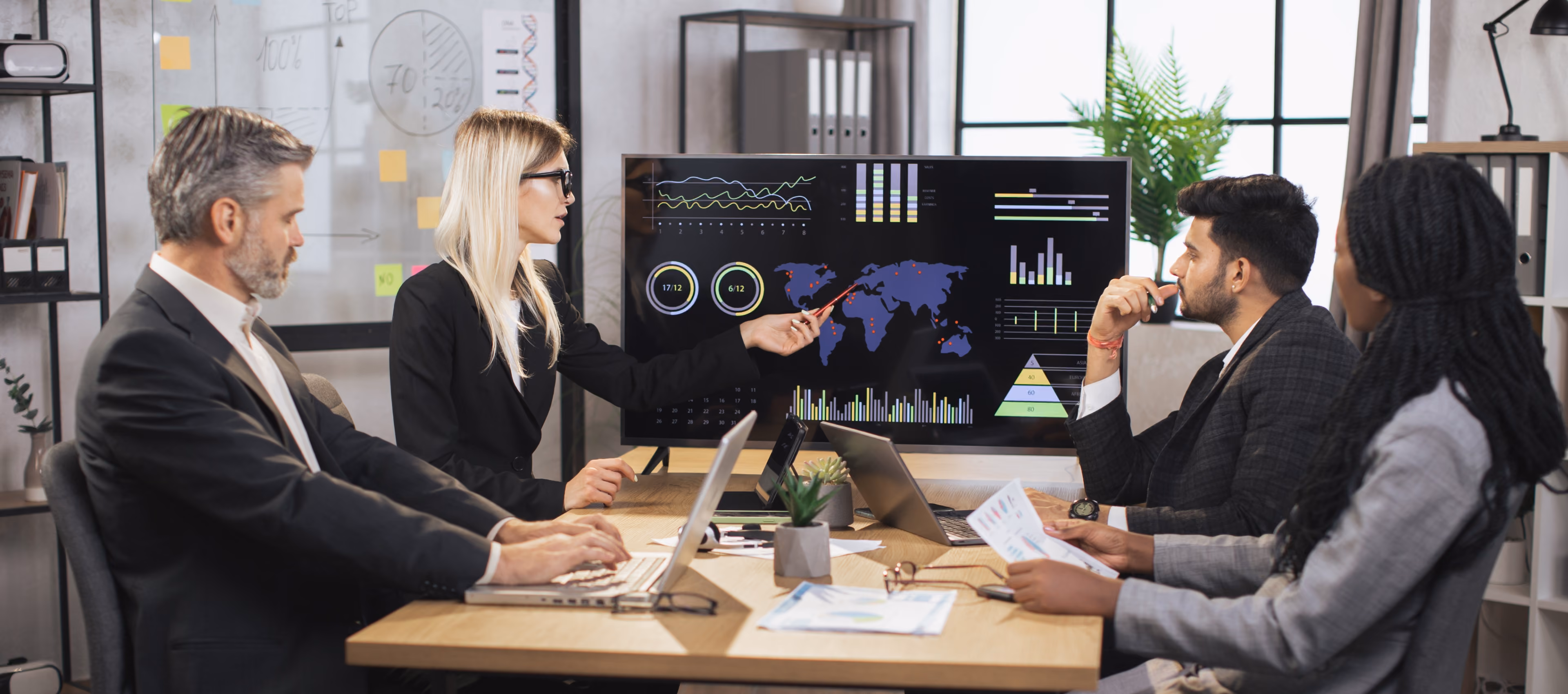 Four business professionals in a meeting room sitting around a table with laptops and papers, one woman pointing at a large screen displaying world maps, charts, and graphs.