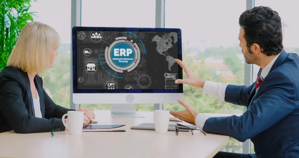 Two business professionals, a woman and a man in suits, sitting at a table with coffee mugs, looking at a computer screen displaying an ERP (Enterprise Resource Planning) interface with icons and a world map.