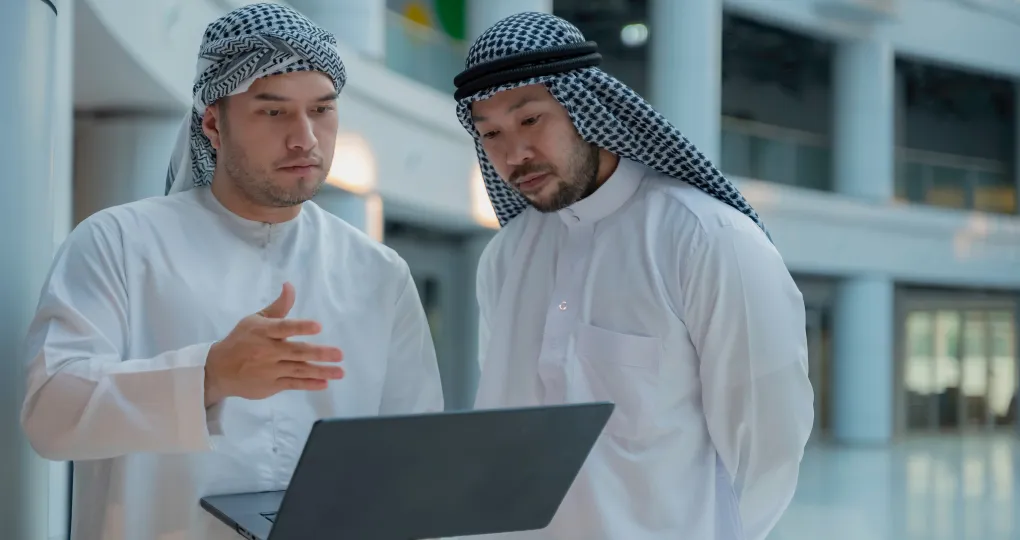 Two men wearing traditional Middle Eastern clothing standing indoors and looking at a laptop screen while one gestures with his hand.