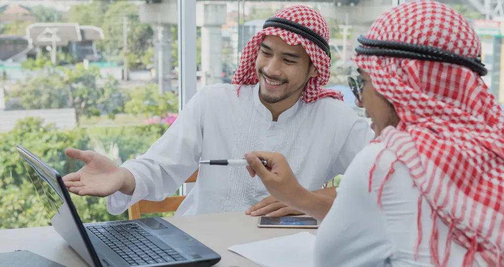 Two men wearing traditional Middle Eastern attire sitting at a table with a laptop, one man smiling and gesturing towards the laptop screen while the other points at it with a pen.