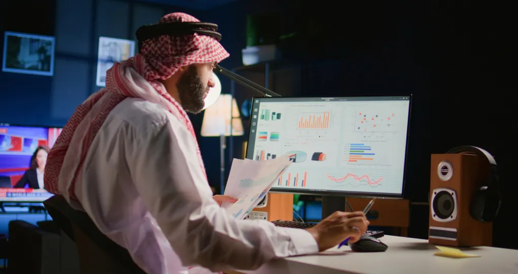 Man in traditional Middle Eastern attire reviewing printed charts and graphs while seated at a desk with a computer monitor displaying various colorful data visualizations and charts in a dimly lit room.