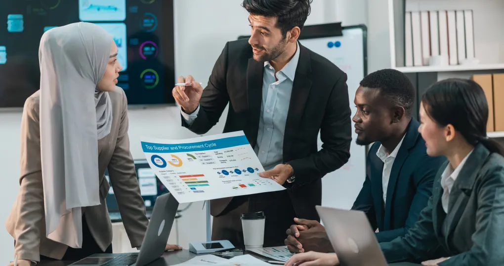 Four diverse business professionals engaged in a meeting around a table with laptops and charts, one man in a suit presenting a paper with graphs and data to the others.