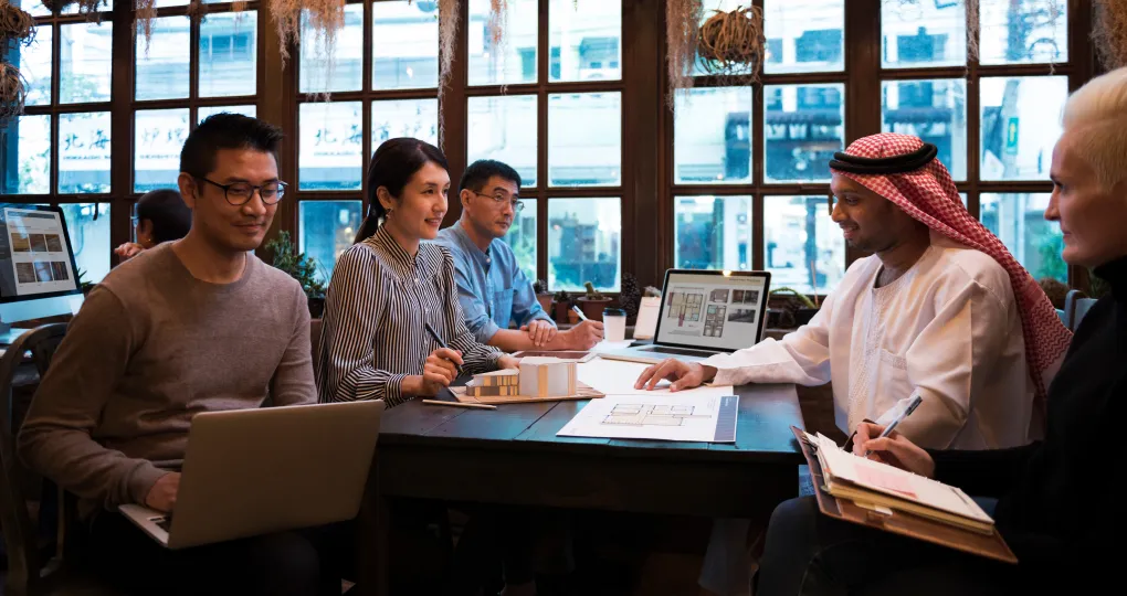 Five people of diverse backgrounds sitting around a table in a bright office with large windows discussing architectural plans and a scale model, with laptops and notebooks open.