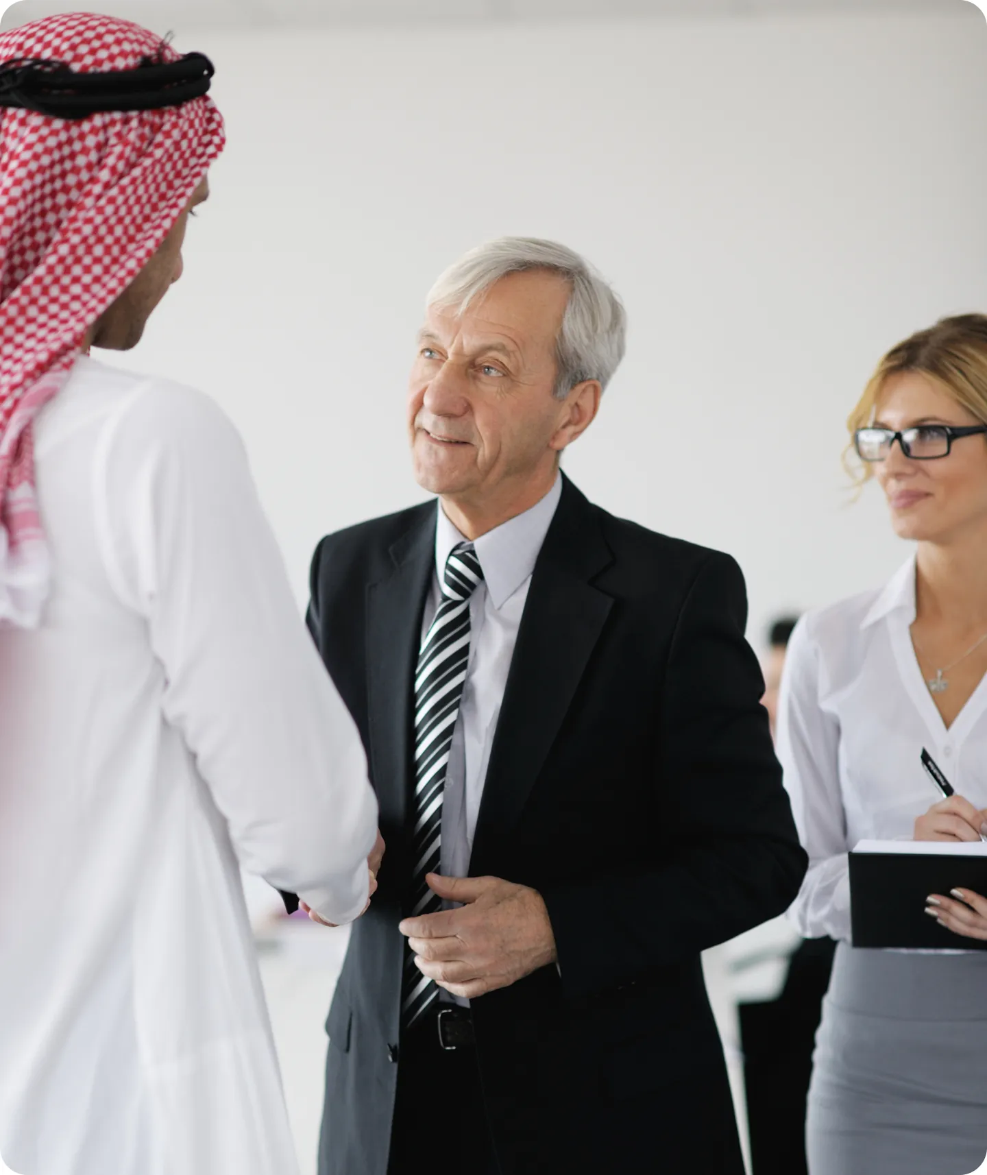 A man in a black suit and striped tie shaking hands with a man in traditional Middle Eastern attire with a red and white checked headscarf, while a woman in glasses and business attire stands nearby holding a notepad and pen.