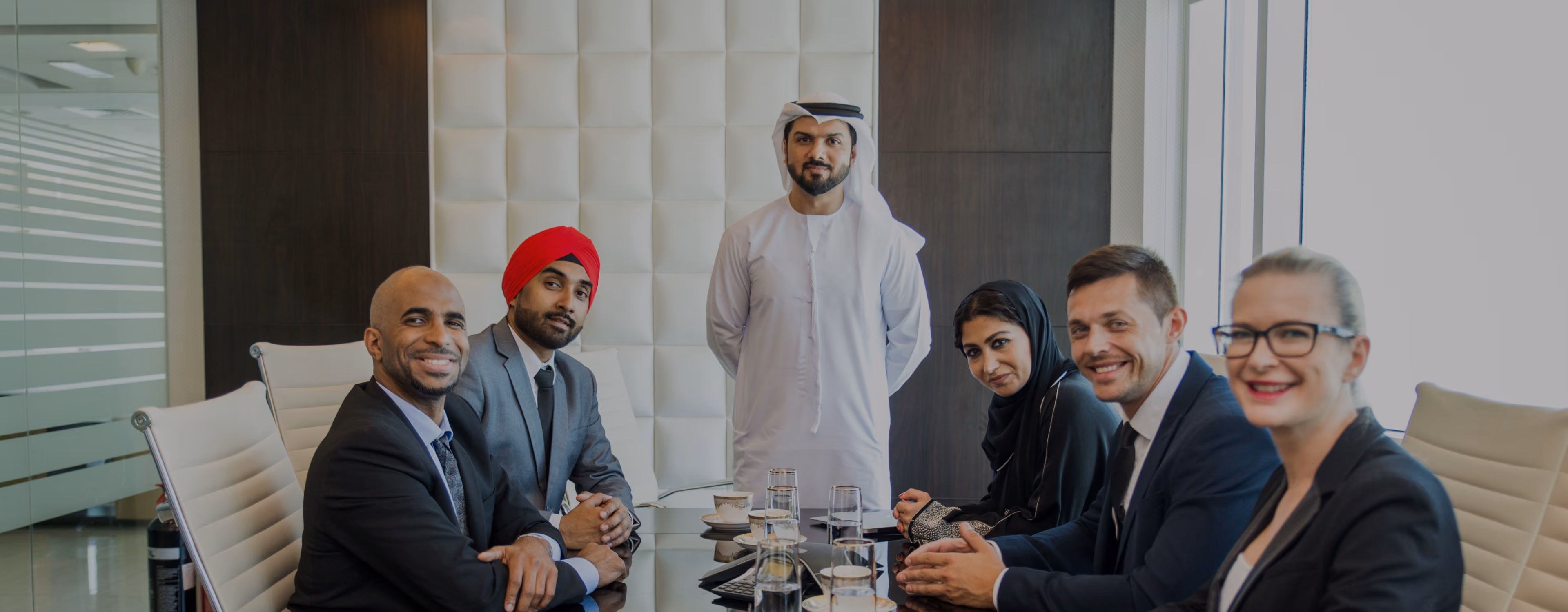A diverse group of six professionals seated around a glass conference table in a modern office, including men and women in business attire and one man standing in traditional Middle Eastern dress.