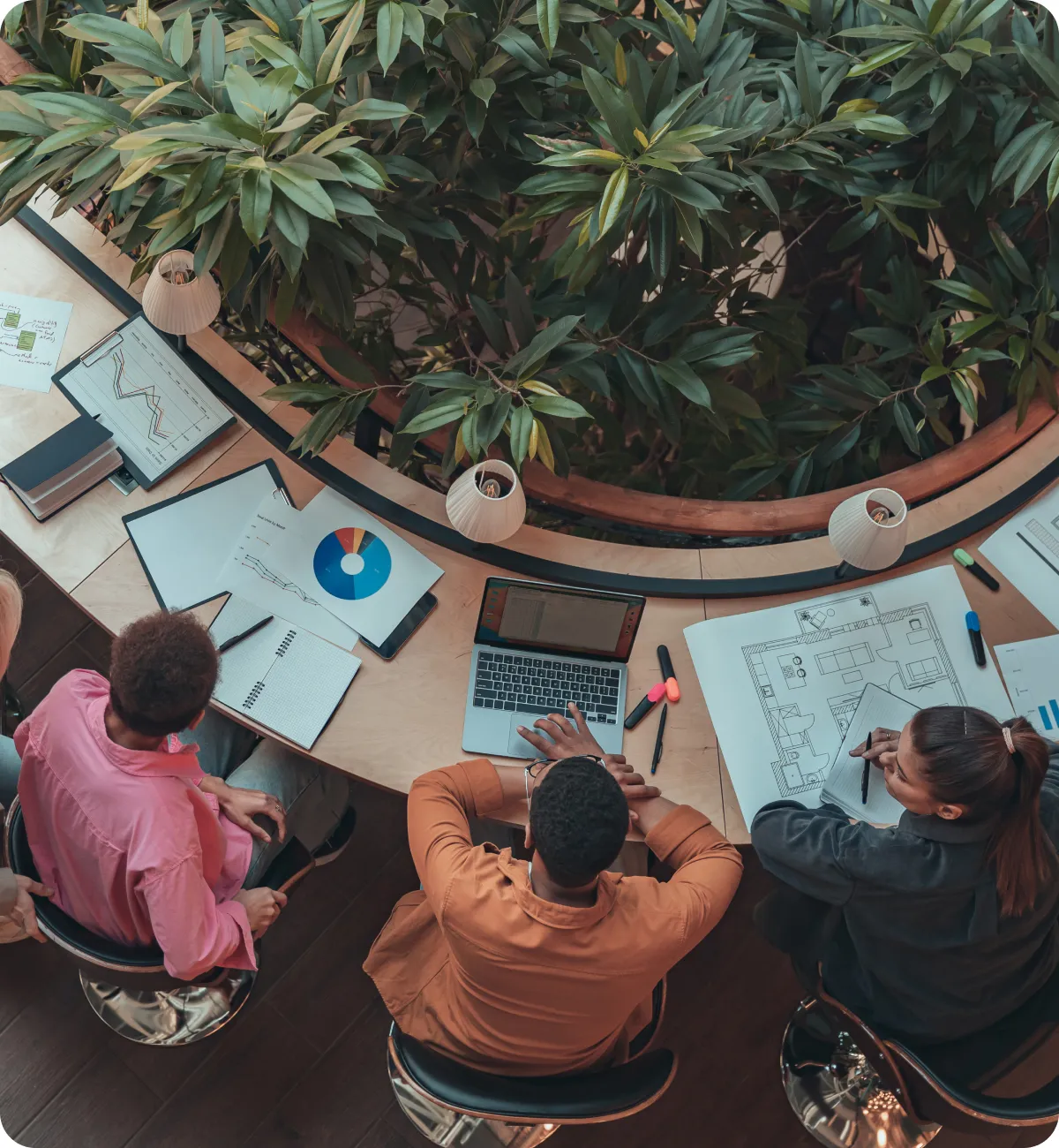 Overhead view of three people seated at a curved wooden desk working with notebooks, charts, a laptop, and architectural plans, surrounded by lush green plants.