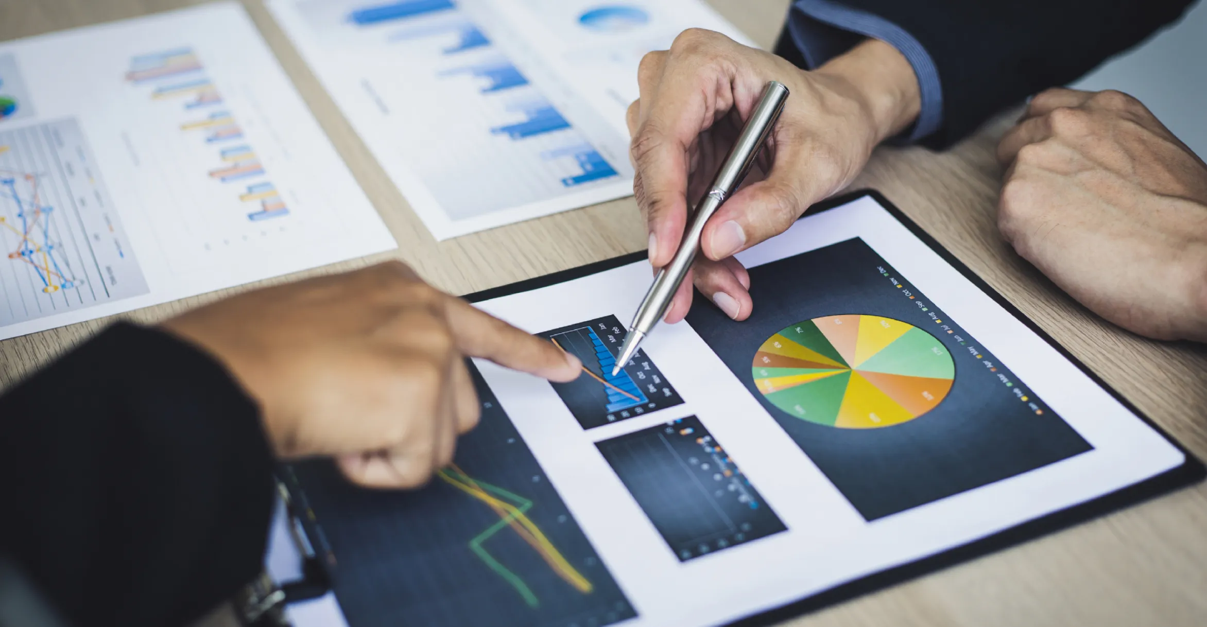 Two people analyzing printed charts on a table, with one person pointing at a bar chart and the other holding a pen near a pie chart and other graphs.