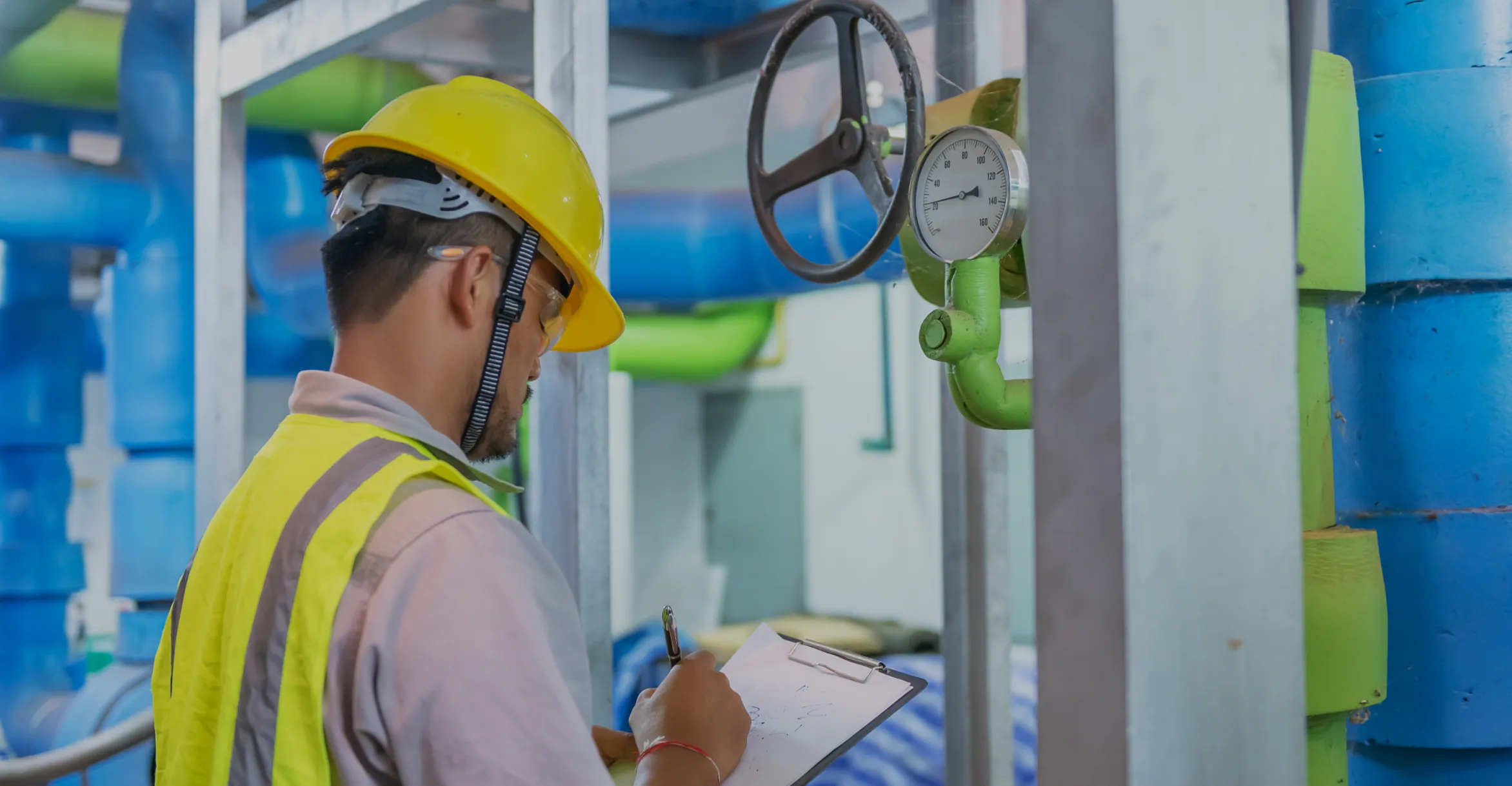 Worker wearing a yellow hard hat and reflective vest inspects and writes on a clipboard in an industrial setting with blue and green pipes and a pressure gauge.