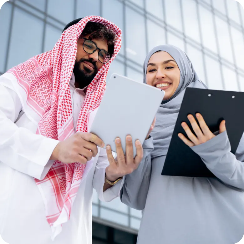Two people dressed in traditional Middle Eastern attire stand outside a modern glass building, looking at a tablet together and smiling, with one holding a clipboard.