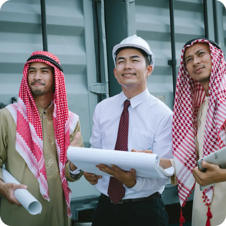 Three men stand together discussing construction plans; two wear red and white keffiyehs and traditional clothing, the third wears a white hard hat, white shirt, and red tie while holding rolled up blueprints.
