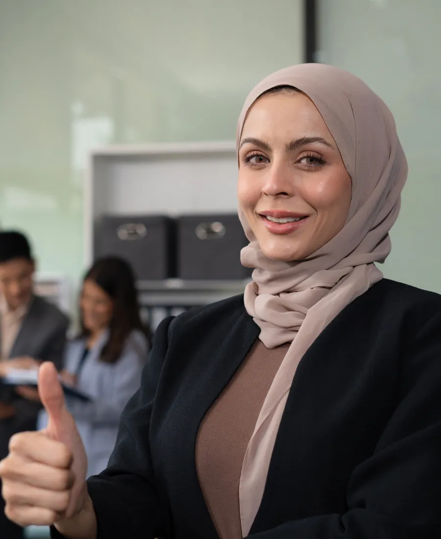 Smiling woman wearing a beige hijab and black blazer giving a thumbs-up in an office setting with two colleagues blurred in the background.