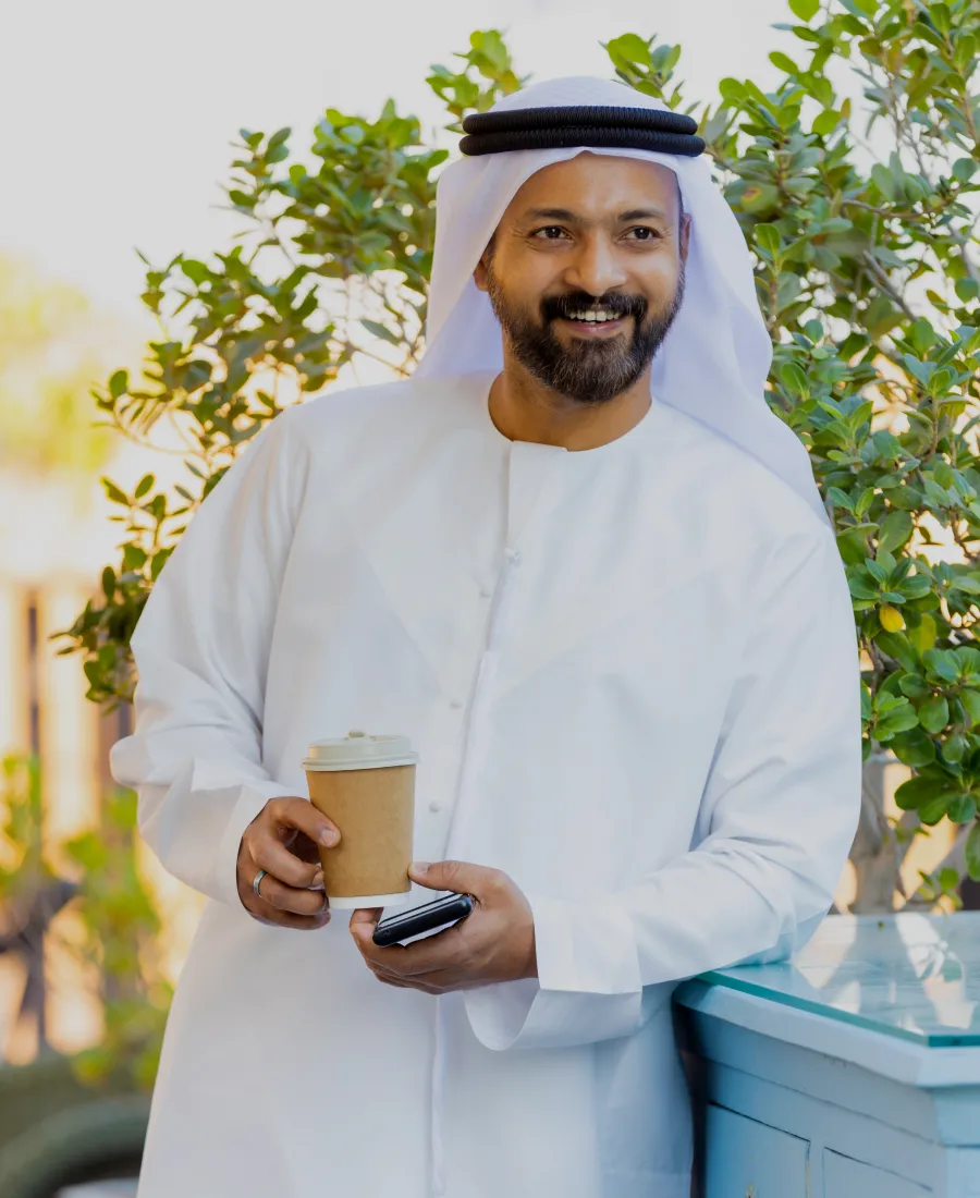 A smiling man wearing a traditional white kandura and headscarf stands outdoors holding a brown disposable coffee cup and a smartphone, with green leafy plants in the background.