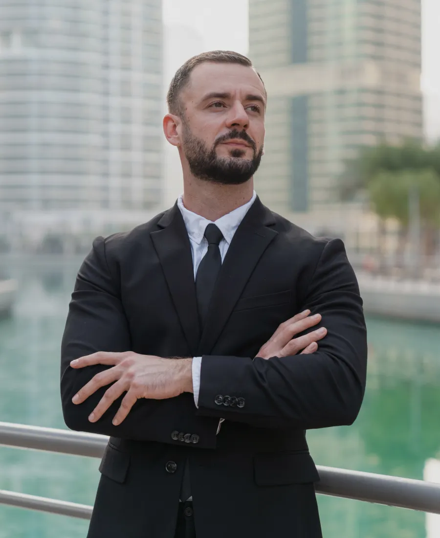 Confident man with beard and short hair wearing a black suit and tie, standing with arms crossed near water and modern buildings in the background.