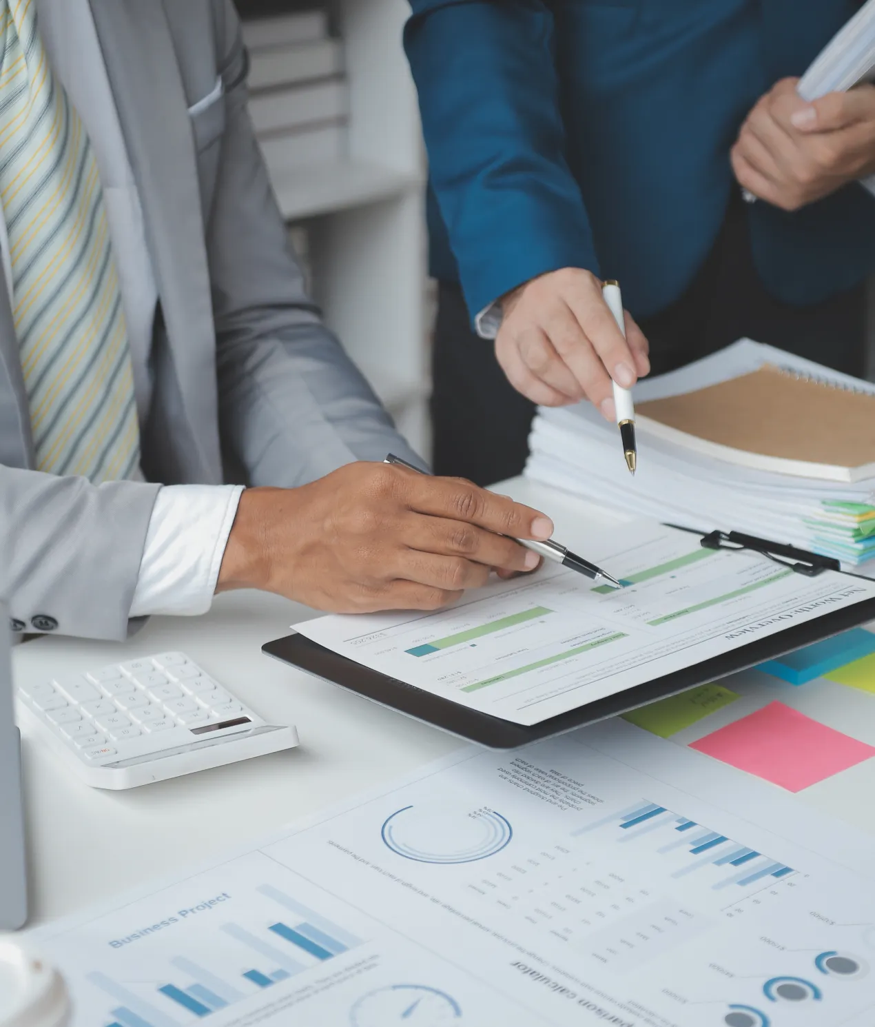 Two business professionals reviewing documents and charts on a desk, one pointing with a pen and the other holding papers, with graphs and a calculator visible on the table.