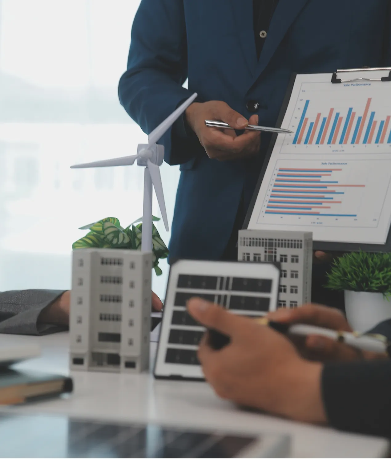 Business person in a blue suit pointing with a pen at a clipboard showing performance bar charts, with a table displaying building models, a wind turbine model, a tablet with solar panel image, and a small potted plant in the foreground.