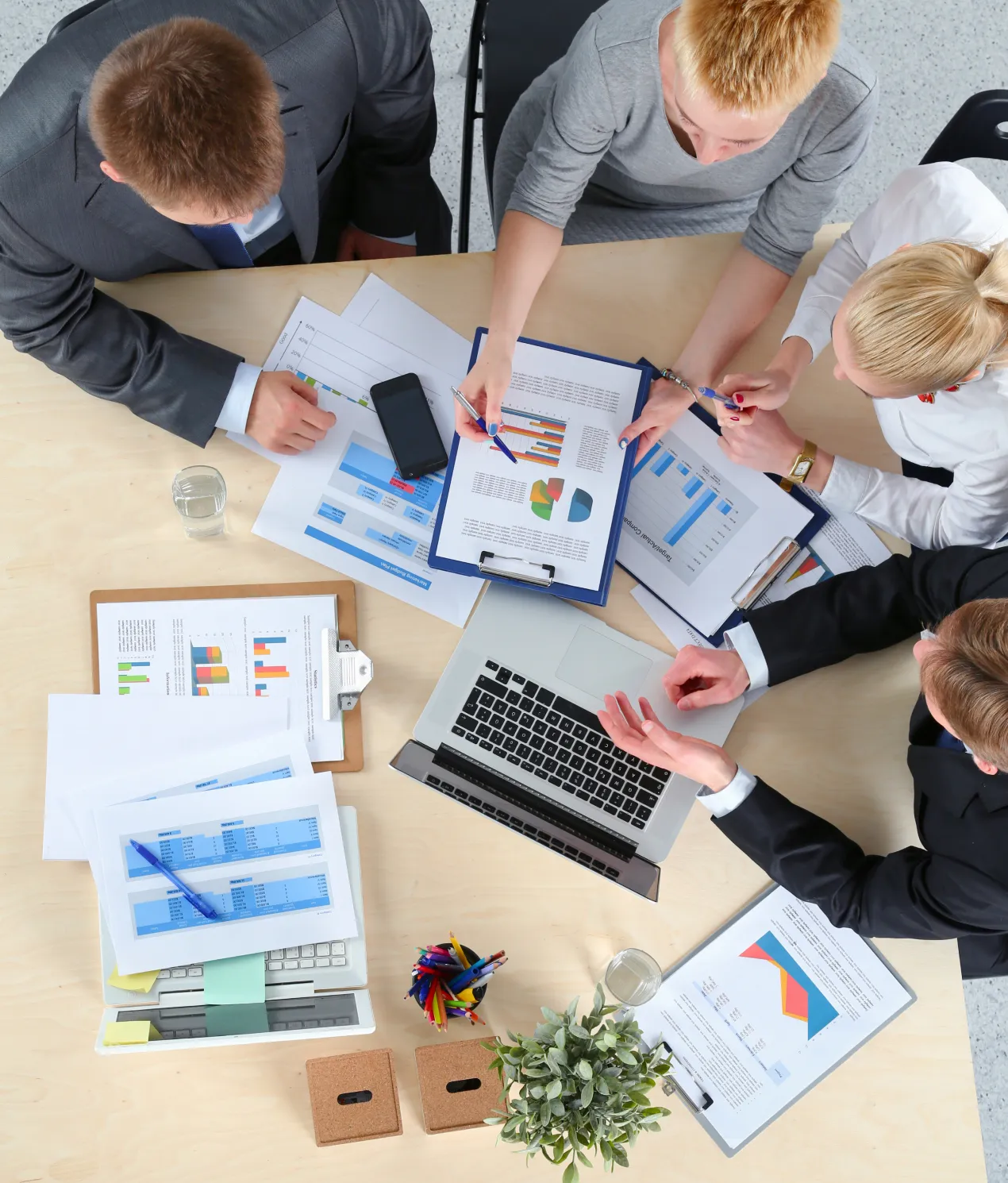 Four business professionals seated around a wooden table reviewing printed charts and reports with colorful graphs, using a laptop and a smartphone, with a small plant and a container of pens on the table.