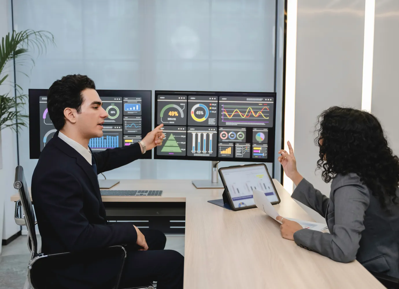 Two business professionals in an office appear to be discussing data displayed on three large monitors with charts and graphs, while a tablet with similar data is on the desk.