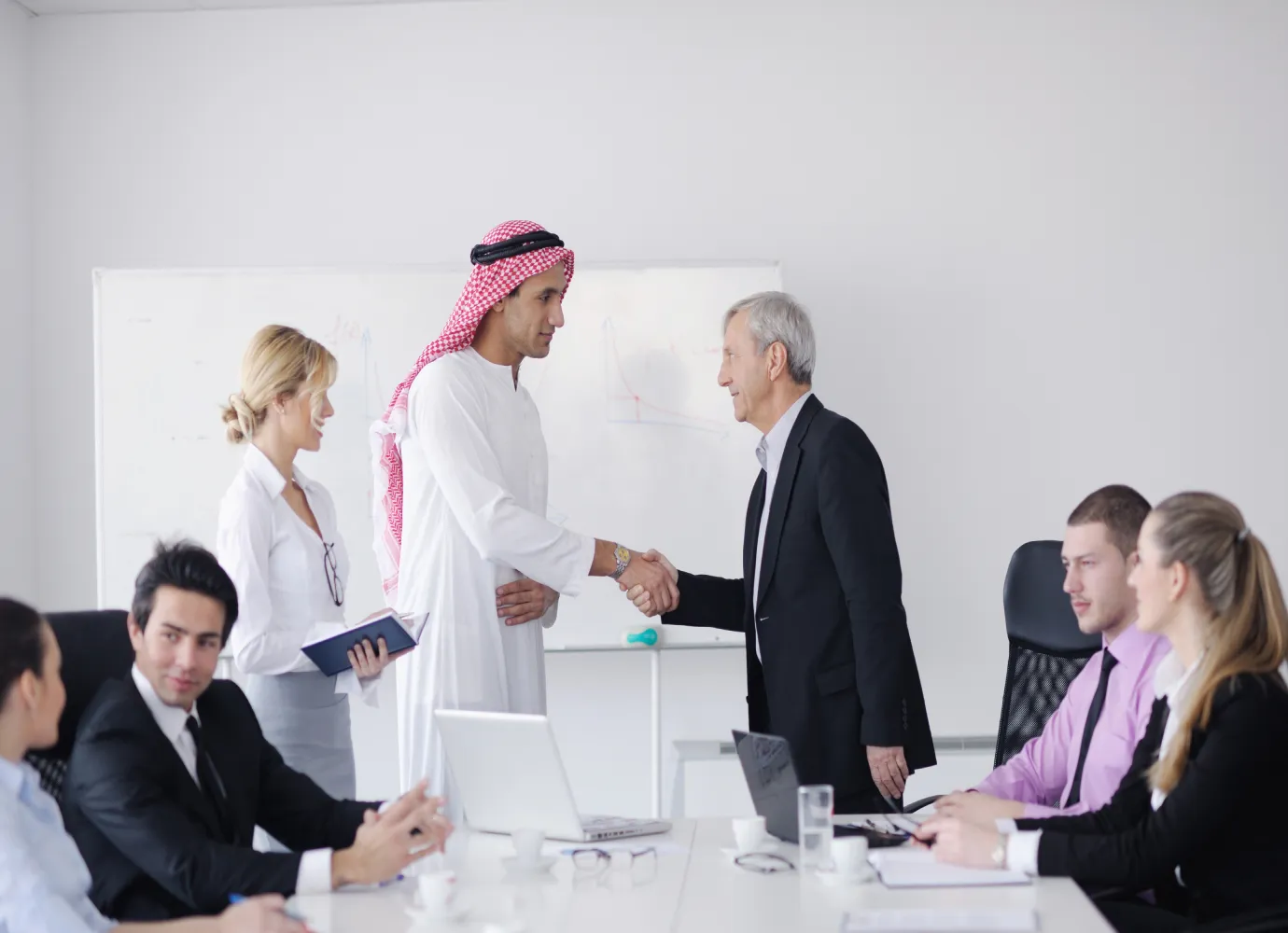 Two men shaking hands in a business meeting room while colleagues sit and observe, with a whiteboard in the background.