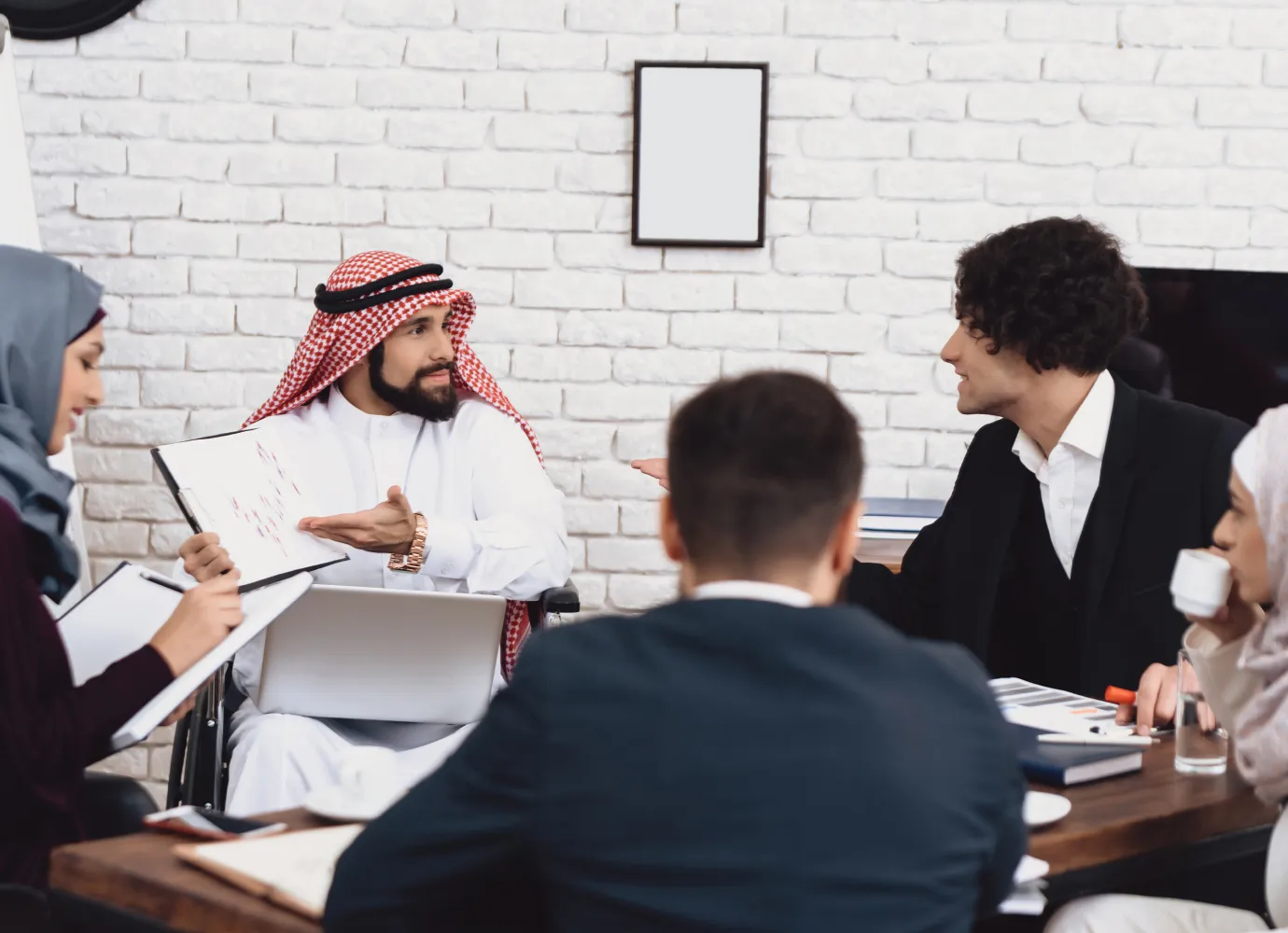 A diverse business meeting with five people, including a man in traditional Middle Eastern attire showing a document, a woman in a hijab taking notes, a man in a suit listening, and another woman drinking coffee, all seated around a table in a modern office with a white brick wall.