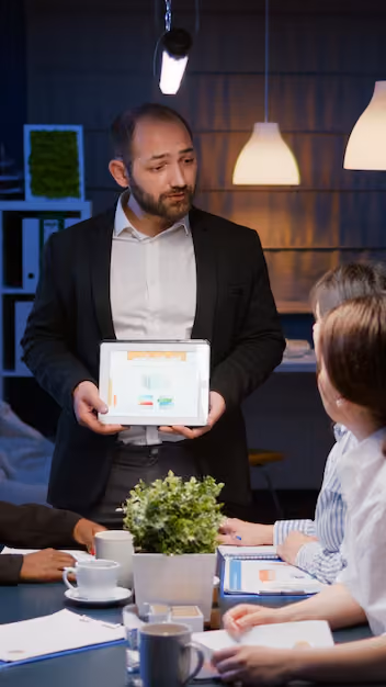 Man in a suit presenting a tablet with charts to colleagues seated around a table in a dimly lit meeting room with modern pendant lights.