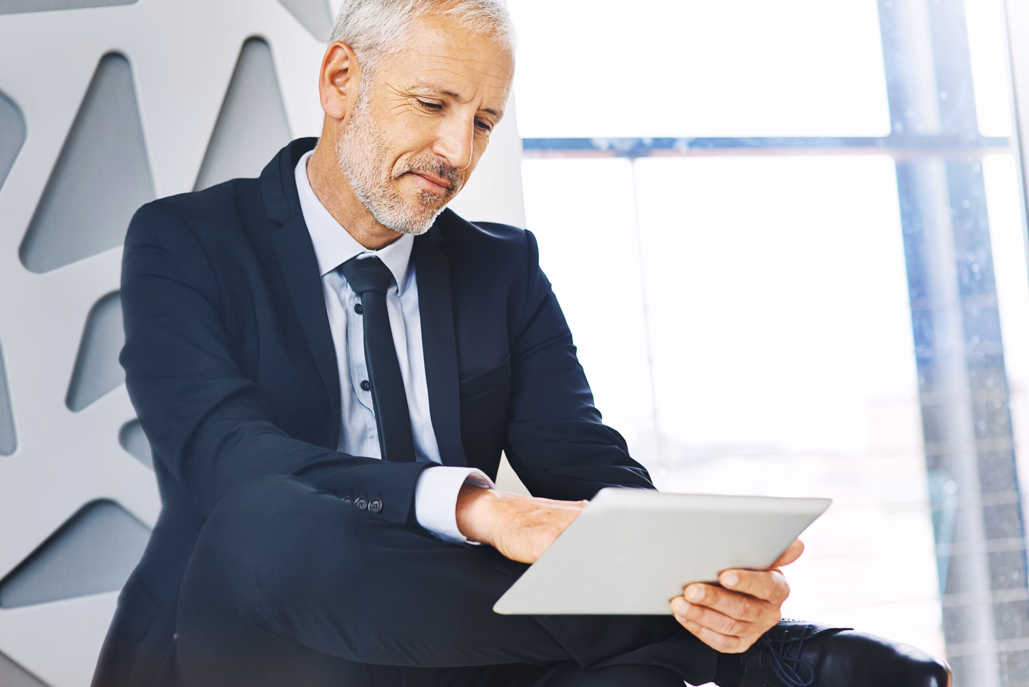 Mature man in a suit sitting and using a tablet device with a focused expression in a modern office setting.