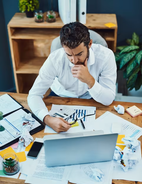 Businessman in a white shirt sitting at a cluttered wooden desk with papers, a laptop, glasses, and a smartphone, looking thoughtful while holding his chin and examining documents.