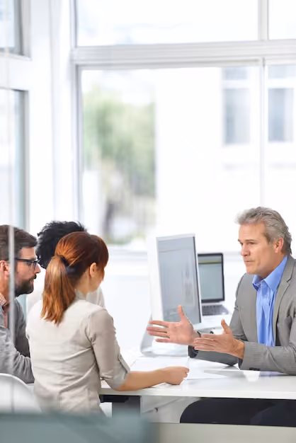 A mature businessman in a gray suit and blue shirt speaks and gestures with his hands across a desk to three younger colleagues in a bright office with large windows.
