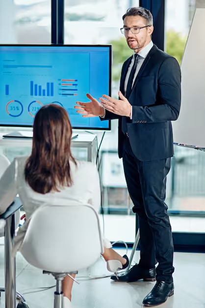 Man in a suit and glasses giving a presentation to a seated woman in a modern office with a large screen showing charts and graphs.