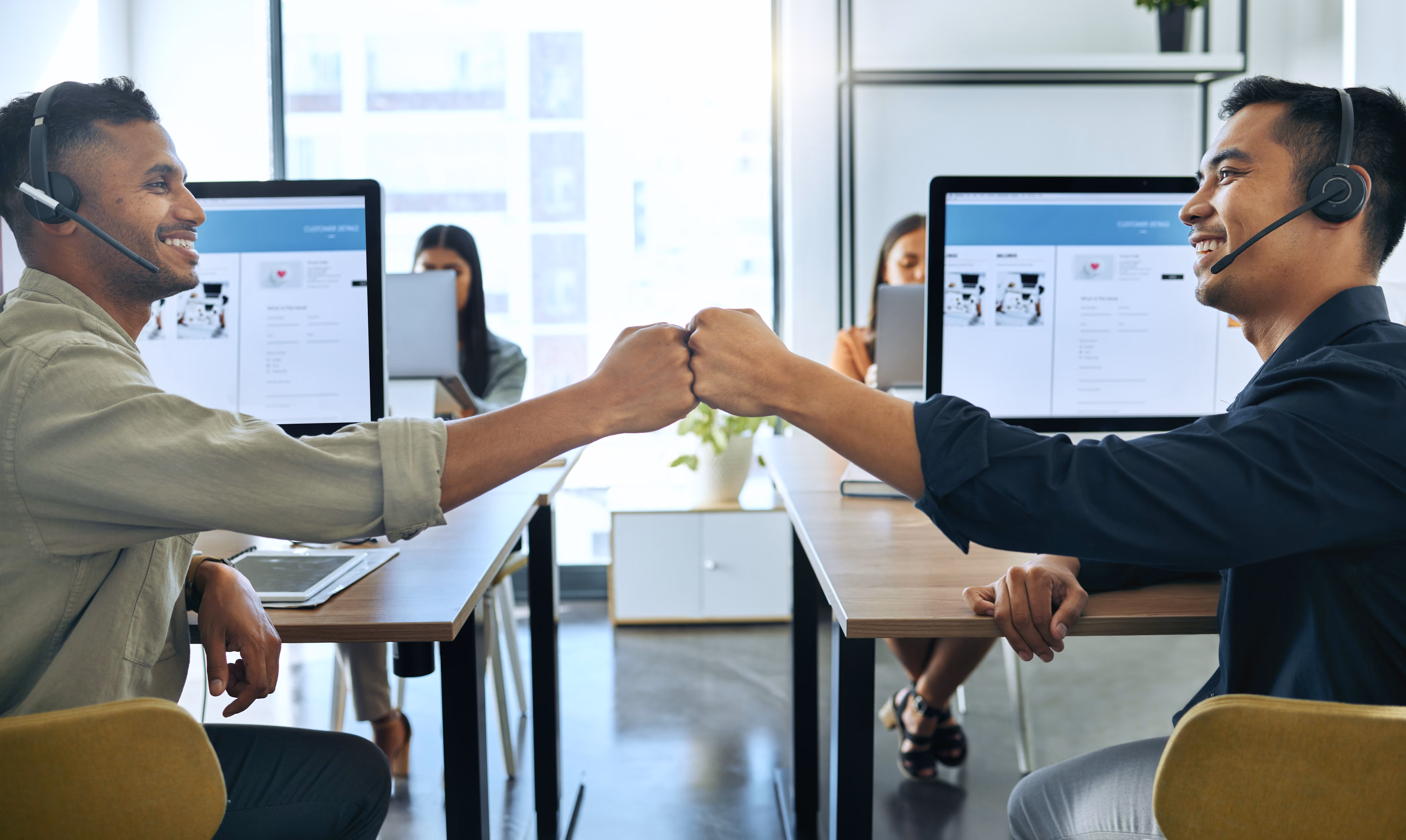 Two male call center employees with headsets sitting opposite each other at desks, smiling and giving a fist bump in a modern office with other workers in the background.