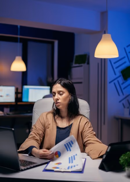Woman in a brown jacket working late at a desk with two laptops and holding papers showing charts and graphs in a dimly lit office with hanging lamps.