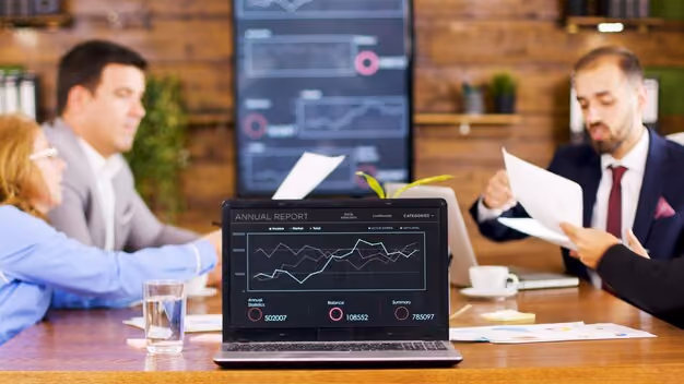 Laptop on a conference table displaying an annual report with graphs and financial data, surrounded by four business people in a meeting.