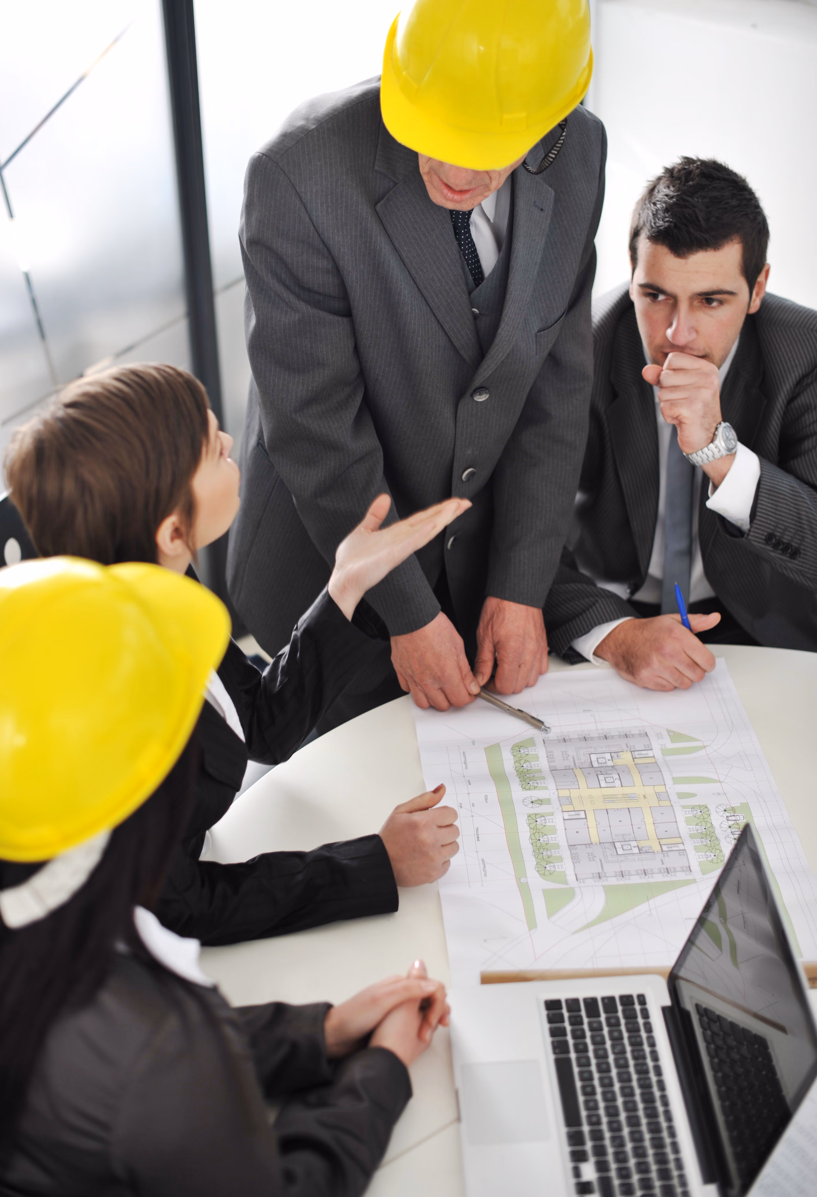 Four business professionals wearing suits, two with yellow safety helmets, gathered around a table discussing architectural blueprints and plans, with one person pointing and others attentively listening.
