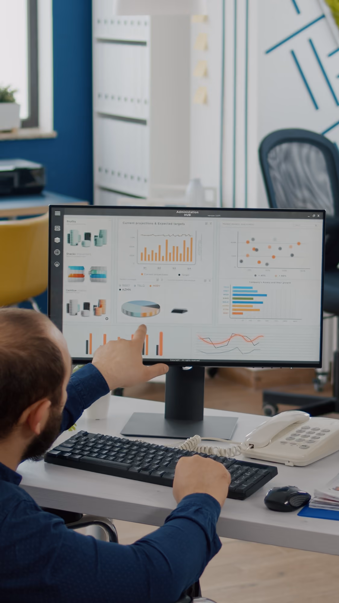Person sitting at a desk pointing at a computer monitor displaying various data charts and graphs in an office environment.