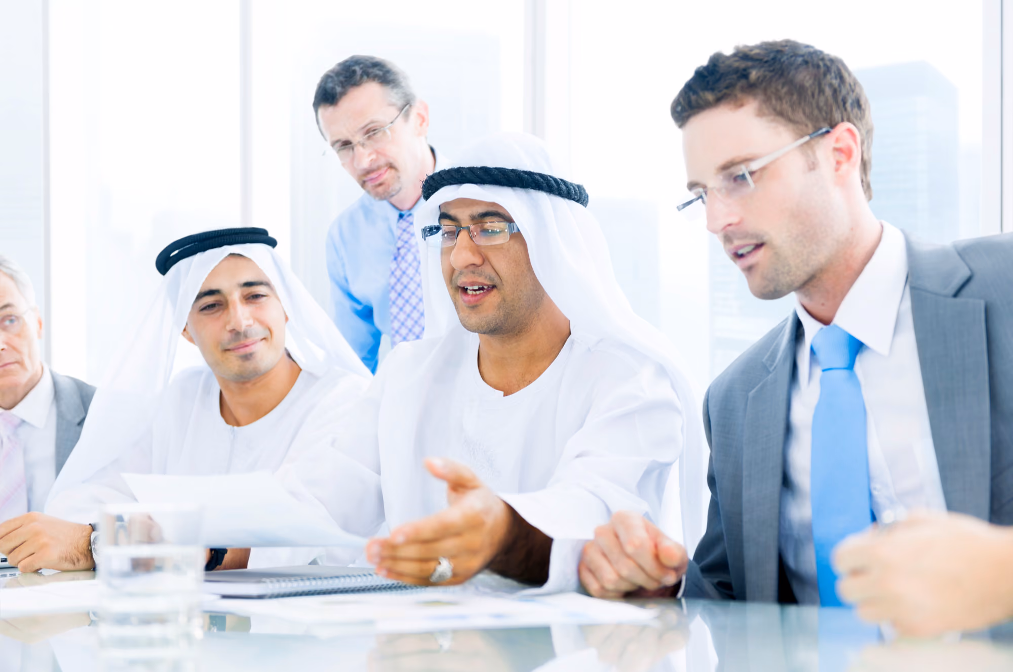 Five business professionals engaged in a discussion around a table in a bright office, including two men in traditional Middle Eastern attire and three men in Western business suits.