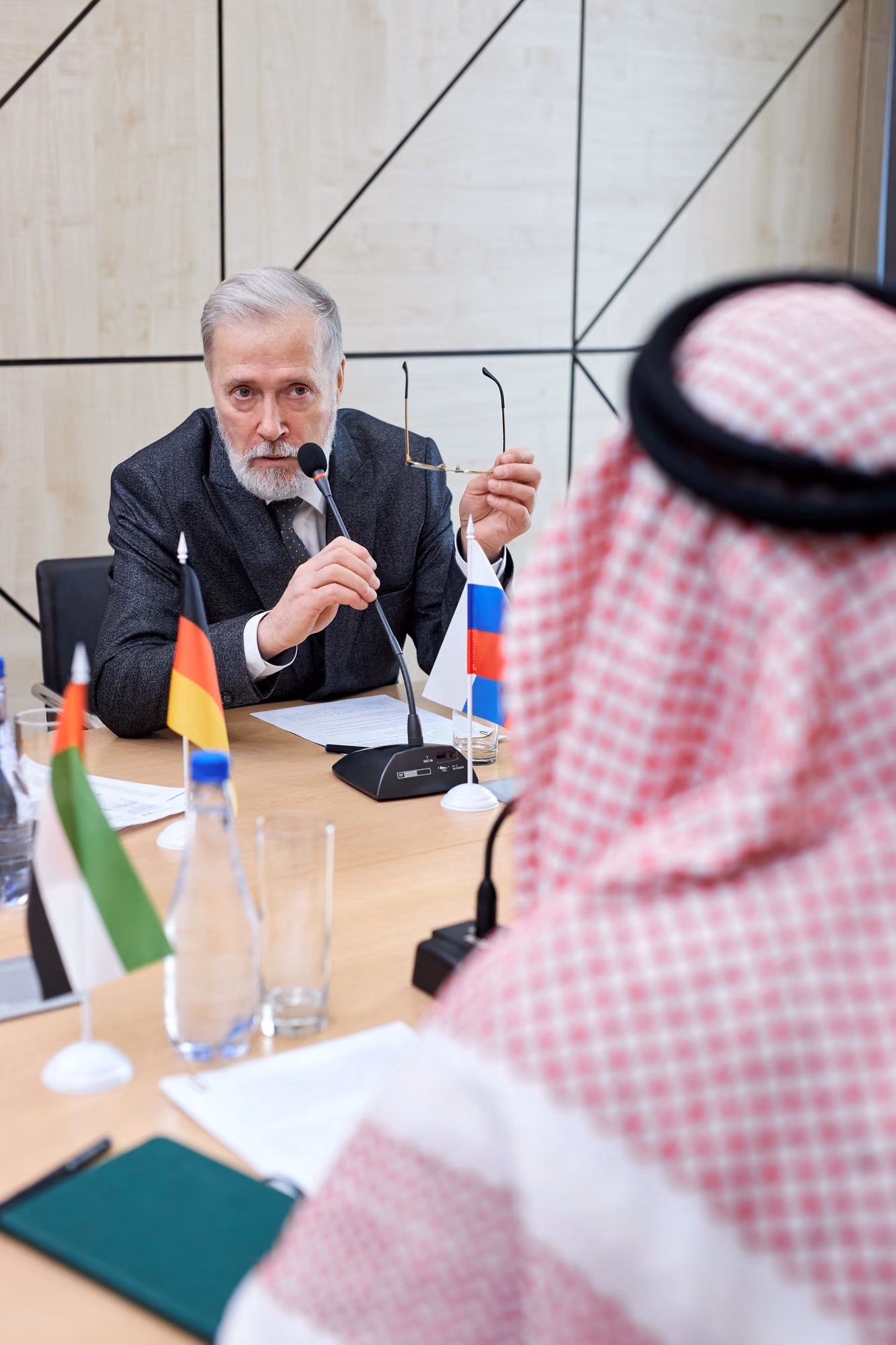 Two men in a formal meeting room, one older man with gray hair and beard speaking into a microphone and holding glasses, the other man wearing traditional Middle Eastern attire, with German, Russian, and United Arab Emirates flags visible on the table.