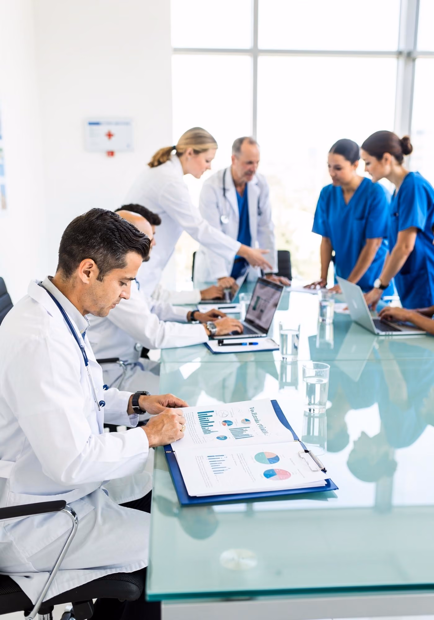 Group of medical professionals, including doctors and nurses, gathered around a glass conference table in a bright room, reviewing charts and working on laptops during a meeting.