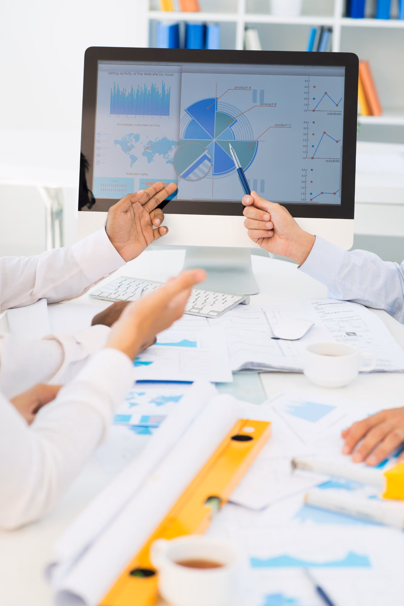 Two people in a business setting pointing at charts and graphs on a computer screen, discussing data and reports on a cluttered desk with papers and cups.