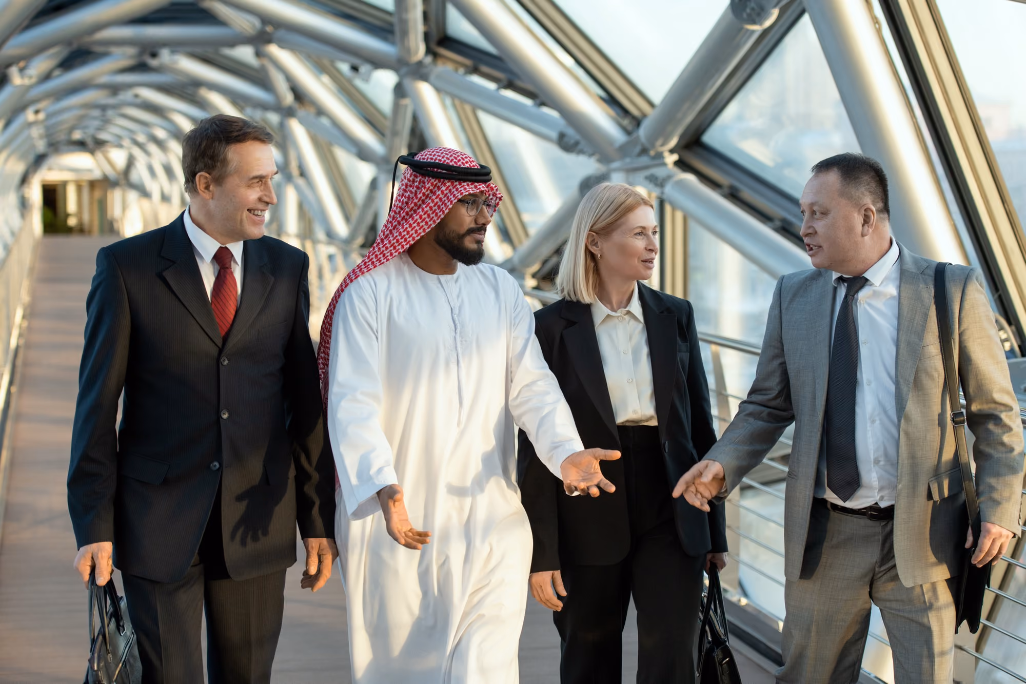 Four business professionals walking and talking together in a modern glass-enclosed walkway, including a man in traditional Middle Eastern attire, two men in suits, and a woman in a black suit.