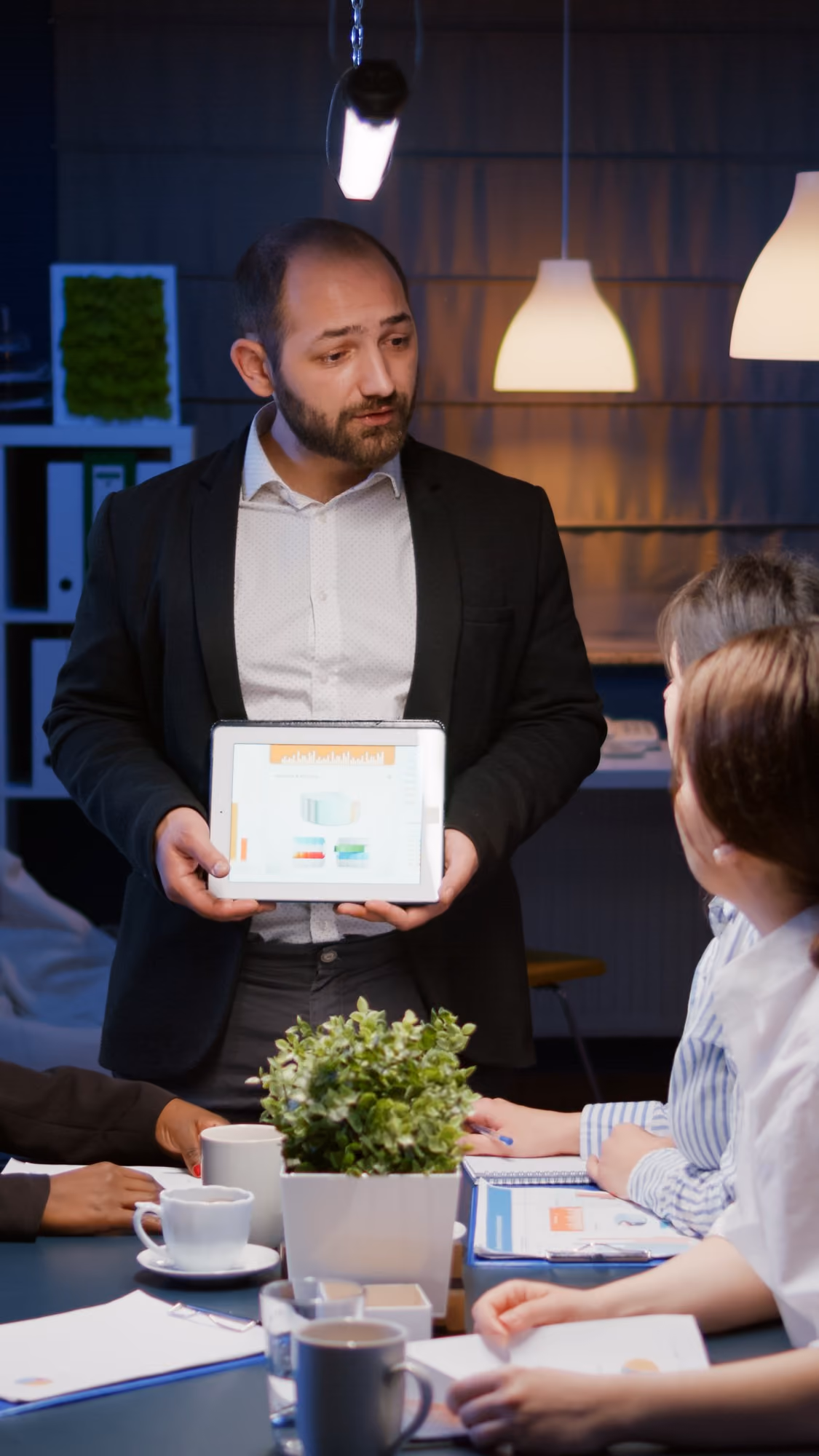 A man in a black blazer and white shirt holding a tablet showing a presentation with charts, speaking to colleagues seated around a table with documents, notebooks, coffee cups, and a potted plant in a modern office setting with pendant lighting.