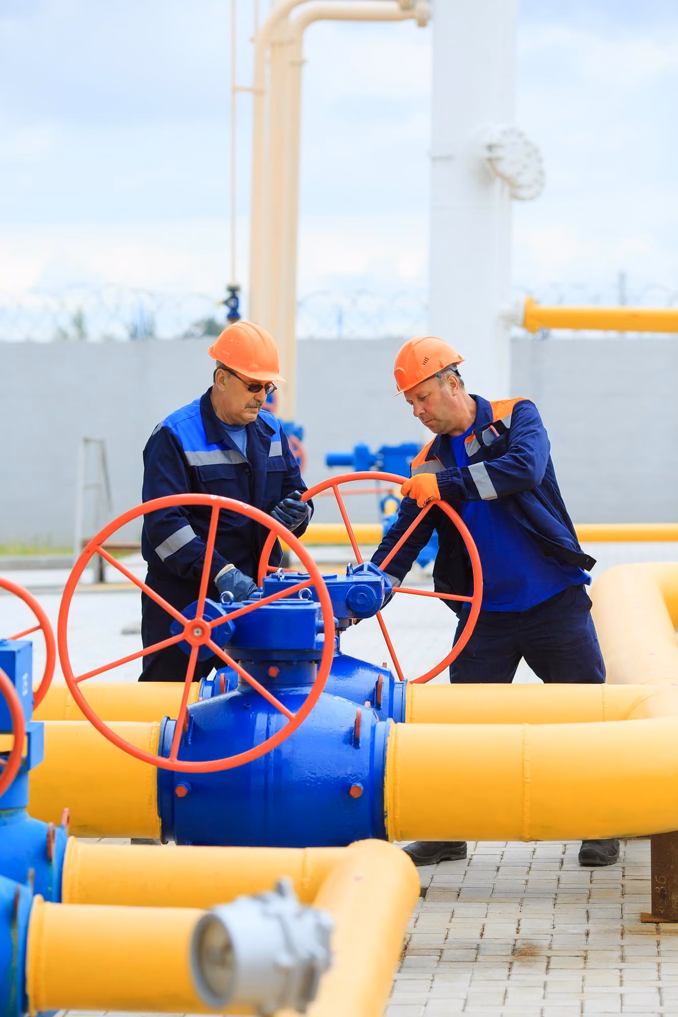 Two workers in safety helmets and uniforms operate large blue valves connected to yellow industrial pipes outdoors.
