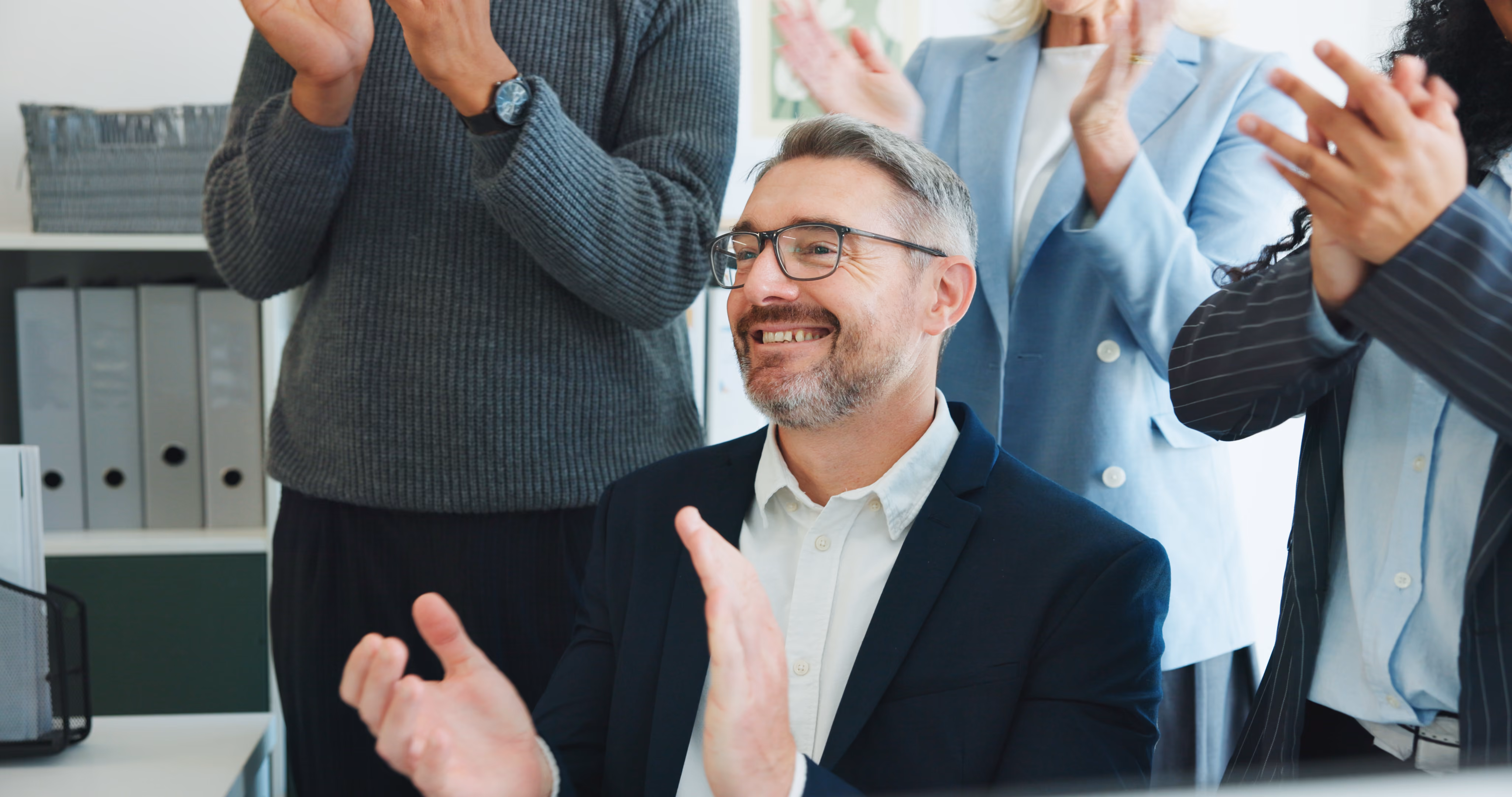 Group of professional colleagues standing and sitting in an office, smiling and applauding a man with glasses wearing a suit.