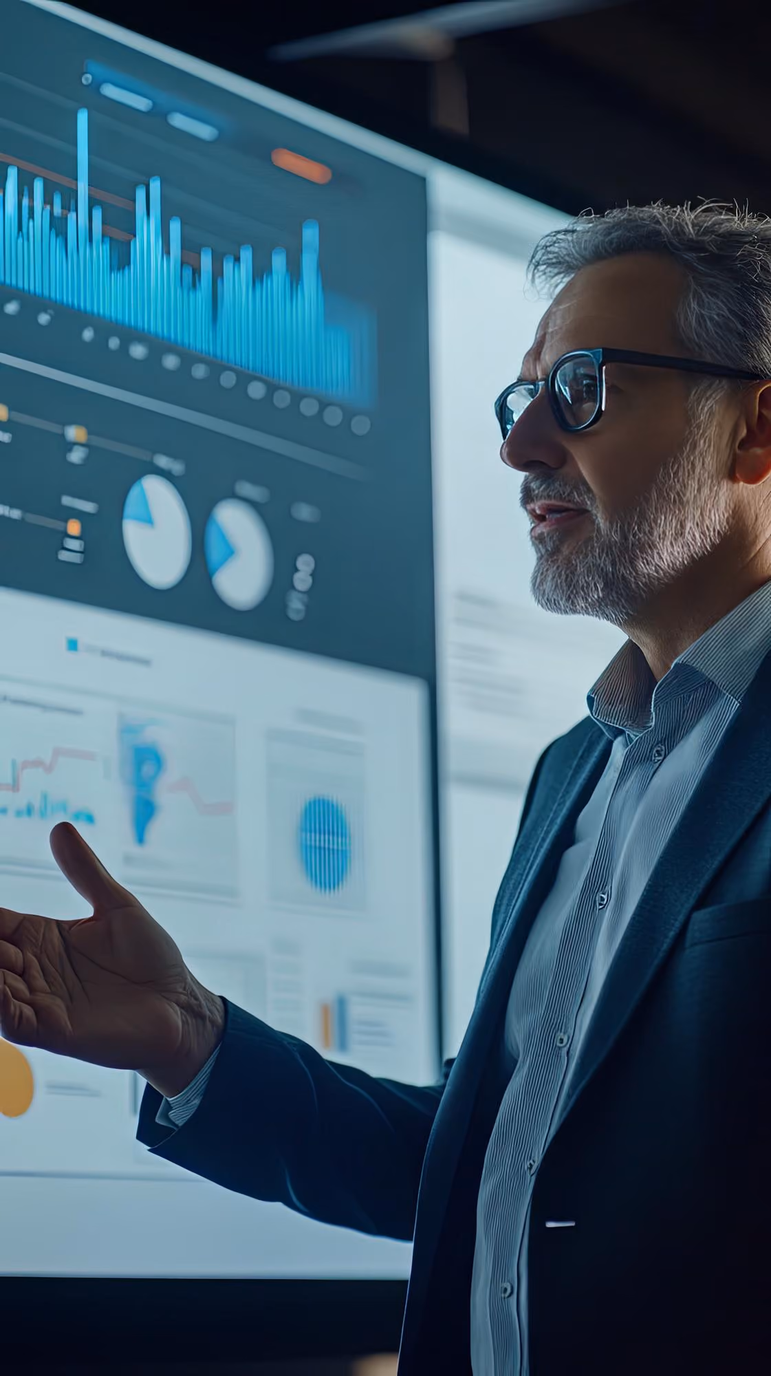 Man in glasses and suit jacket gesturing toward a large screen displaying colorful data charts including bar graphs and pie charts in an office setting.