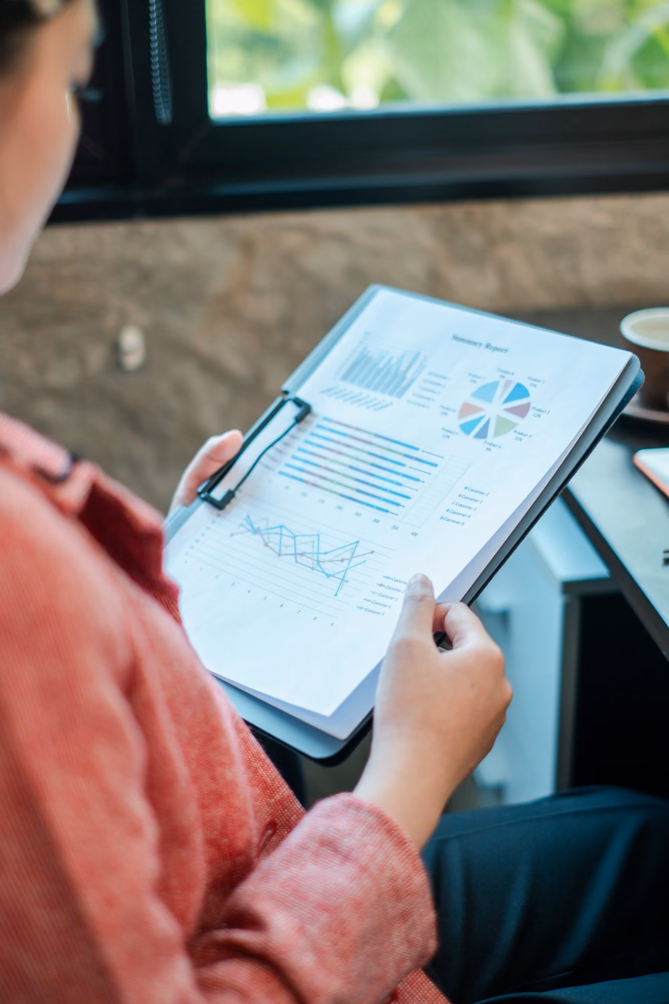 Person in a red sweater holding a clipboard with various charts and graphs, including bar charts, a pie chart, and a line graph, seated near a window with greenery outside.
