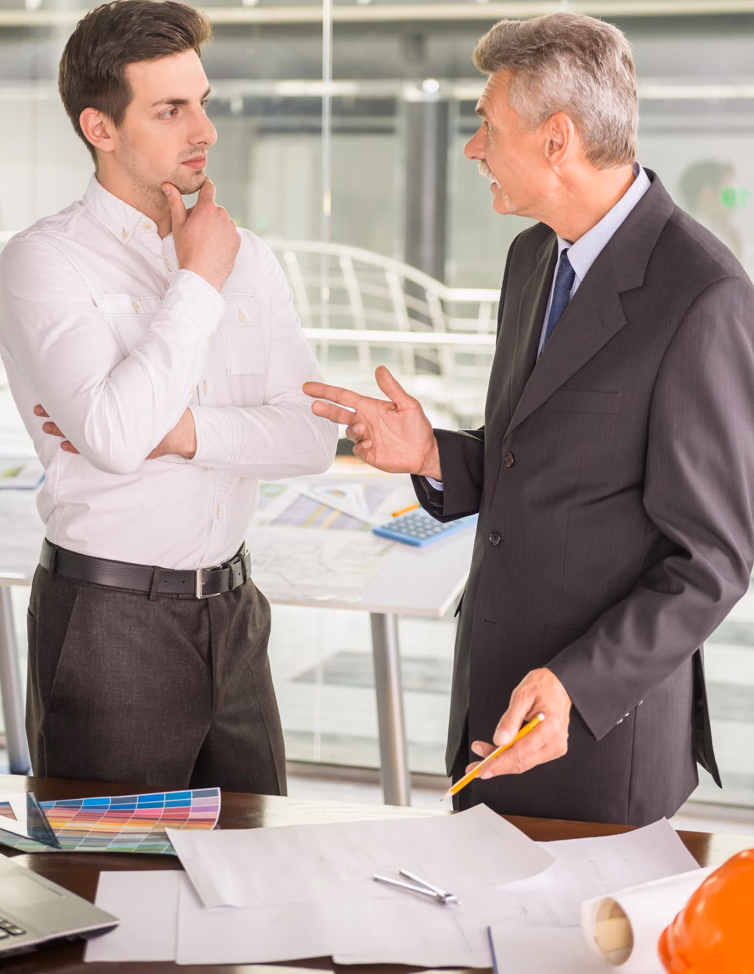 Two men in an office discussing plans; one in a white shirt thoughtfully listens with hand on chin, the other in a dark suit gestures with a pencil over paperwork and architectural tools on the table.