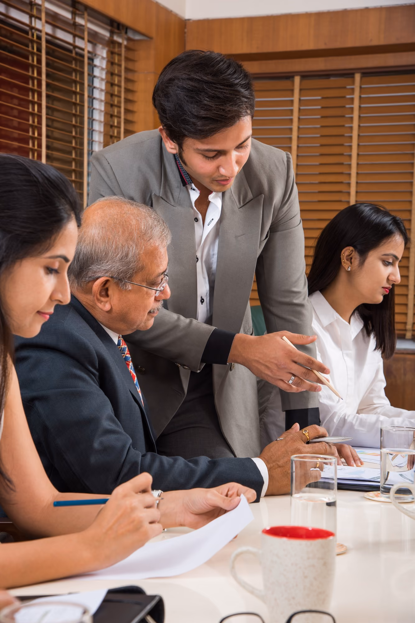 Four professionals in a meeting room with wooden blinds; a man in a gray suit leans over the table explaining something to an older man in a dark suit, while two women dressed in business attire take notes and review documents.