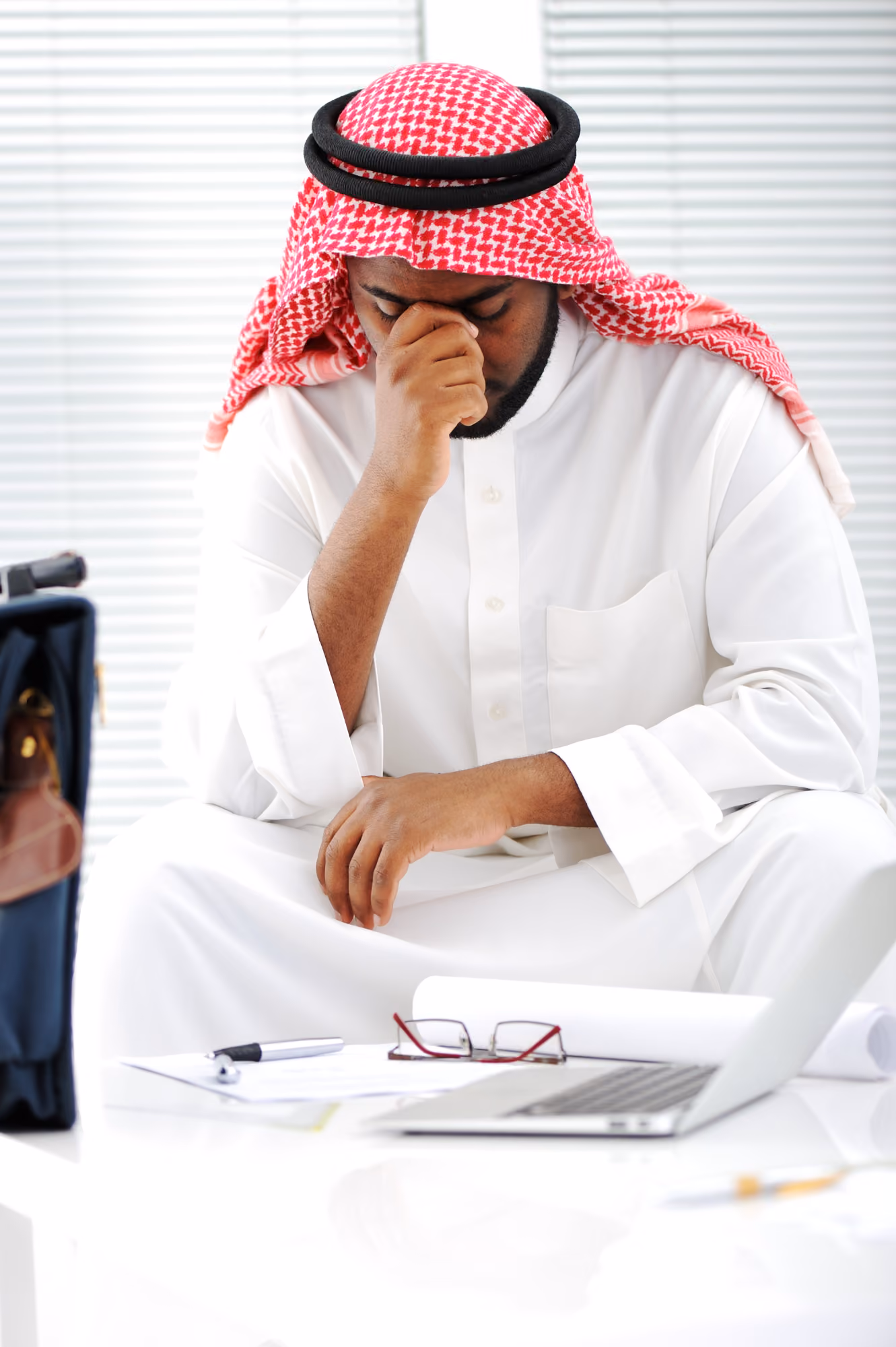 Man wearing traditional Middle Eastern attire sitting at a desk with his head bowed and hand covering part of his face, appearing stressed or contemplative, with glasses, a pen, paper, and an open laptop on the desk.