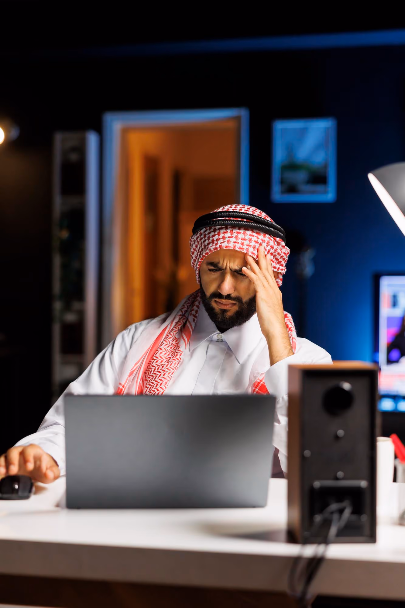A man wearing traditional Middle Eastern attire sits at a desk, looking stressed or confused while working on a laptop, with his hand on his forehead and a computer mouse in the other hand.